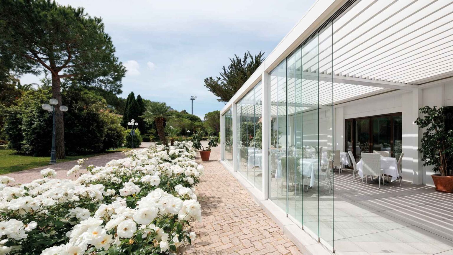Pergola mit Glas-Schiebeanlagen entlang eines Gartens mit blühenden weißen Rosen